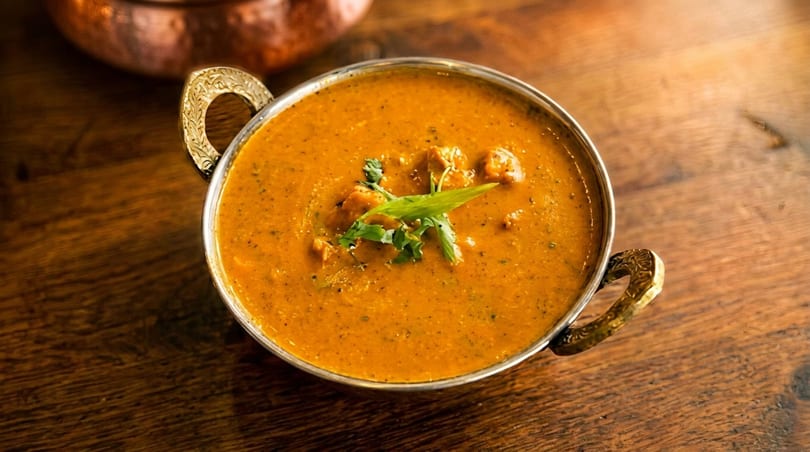 A metal bowl of orange curry with chicken and green herbs sits on a wooden table.