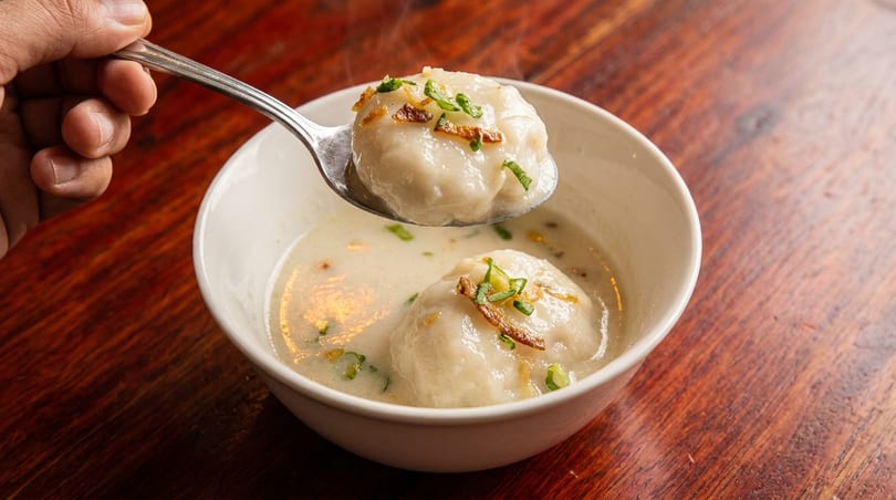 A hand holds a spoon lifting a dumpling from a bowl of creamy soup with green onions.