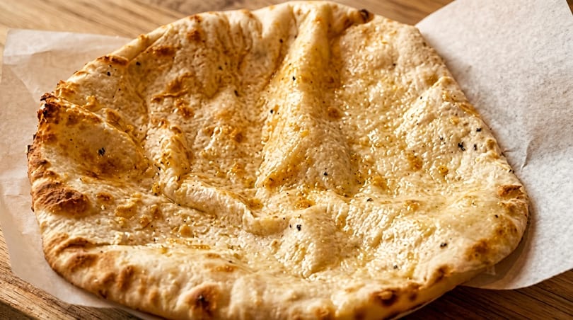 Close-up of a golden-brown naan bread, glistening with butter, on white parchment paper.