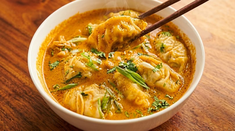 Chopsticks lifting a steaming dumpling from a bowl of orange curry with cilantro and scallions.